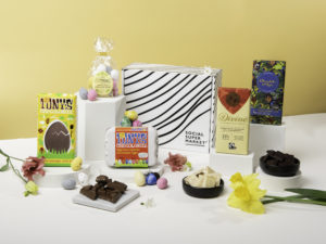 The Eco Easter Bundle Gift Box with its contents around the box they come in, including Tony's chocolate bar, Tony's Easter mini eggs, Divine Chocolate mini speckled eggs, Divine Chocolate white chocolate bar and a bar of Chocolate and Love. There's also daffodils another flowers around the products.