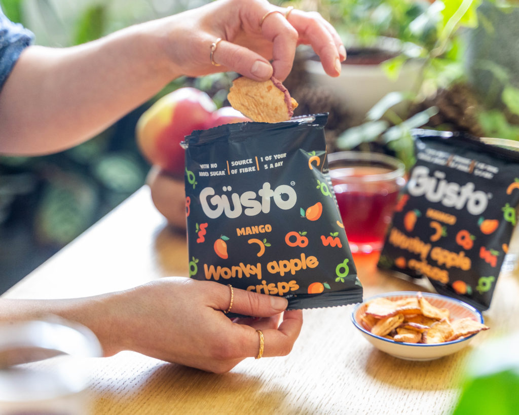 A hand reaches into a bag of Güsto Snacks and pulls out an apple and mango crisp. There's more in the background in a kitchen scene.
