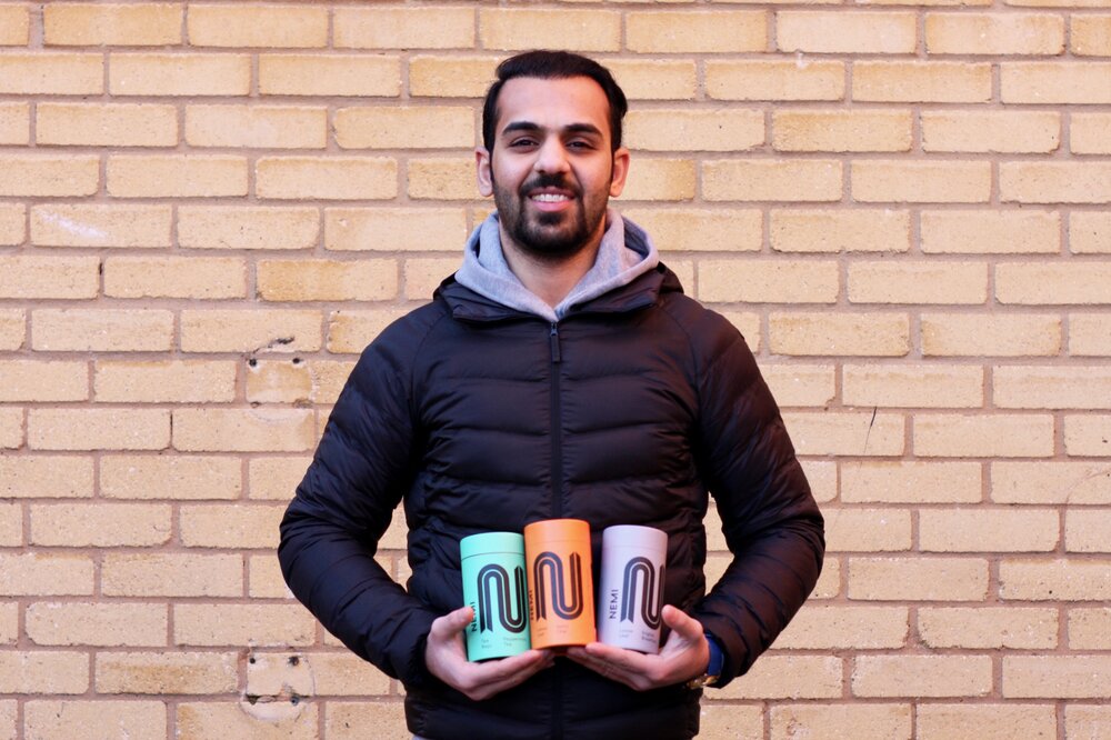 An employee of NEMI Teas holding up three tubes of the tea. He stands smiling against a brick wall