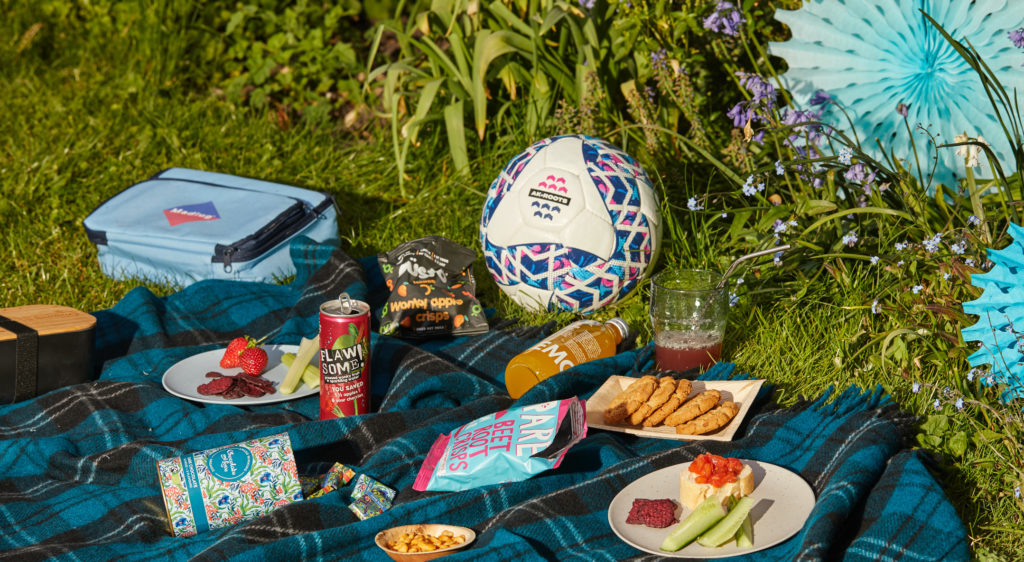 A picnic scene with a Madlug cool bag, an Alive & Kicking Football and snacks on the ground
