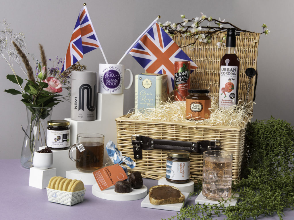 The Best of British Jubilee Hamper with its lid open and contents around it including NEMI Teas, Urban Cordial, Flawsome! drinks, Roots & Wings marmalade, Mama Buci honey, Refuge Chocolate melts and more. The hamper is next to a plant and there's a jubilee commemorative mug with Union Jack flags in.