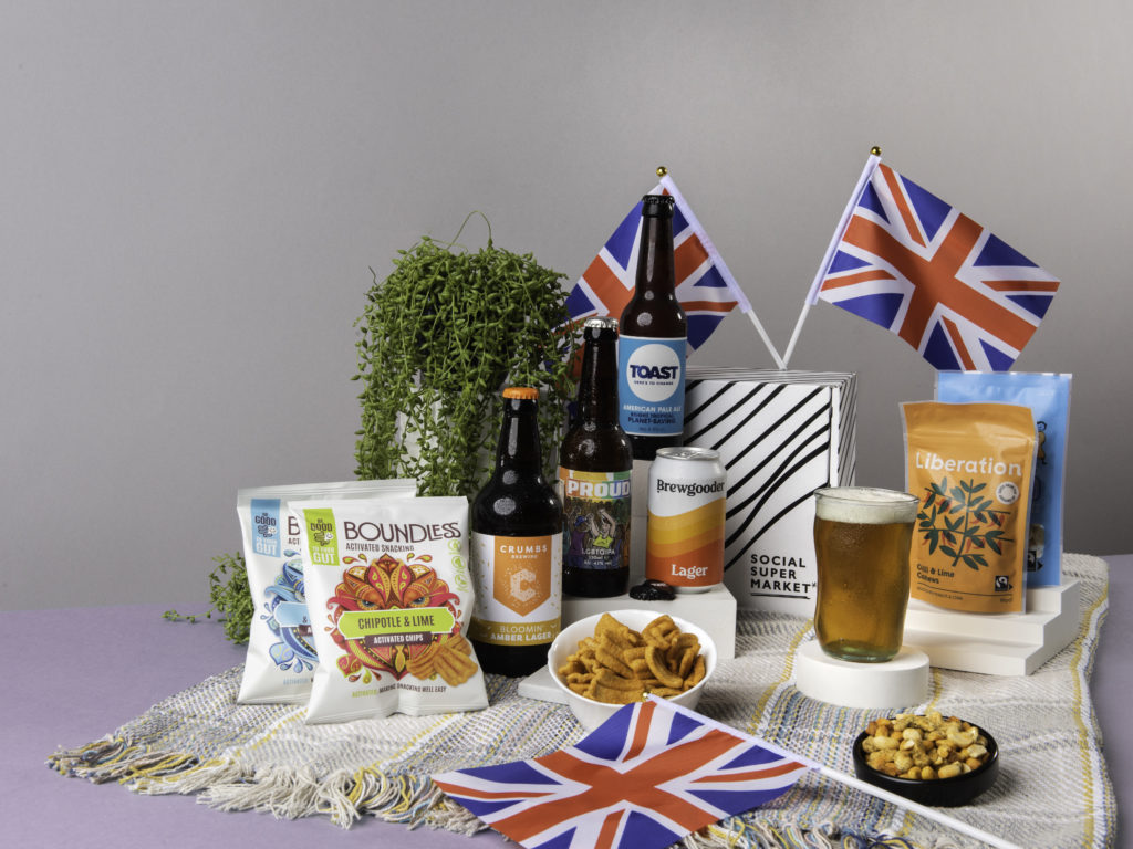 The Jubilee Picnic Beer Tasting Gift Box, surrounded by its content including beers from Crumbs Brewing, Toast Ale, Brewgooder and snacks from Liberation Nuts and Boundless Activated Snacking. They're on a picnic blanket and there's Union Jack flags dotted around them too.