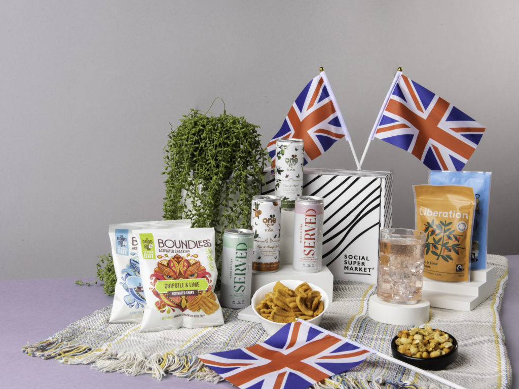 The Jubilee Picnic Gin & Seltzer Tasting Gift Box, surrounded by its content including G&T cans from One Gin and hard seltzers from Served and snacks from Liberation Nuts and Boundless Activated Snacking. They're on a picnic blanket and there's Union Jack flags dotted around them too.