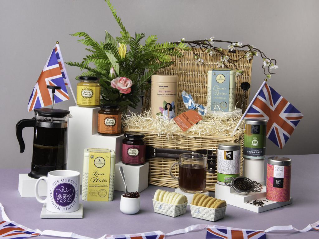 The Jubilee Tea & Biscuits Hamper with its lid open and contents shown around it including preserves from Roots & Wings Organic, teas from Rare Tea Company and coffee from Little Coffee Company. There's also a Platinum Jubilee commemorative mug and Union Jack flags around the products.