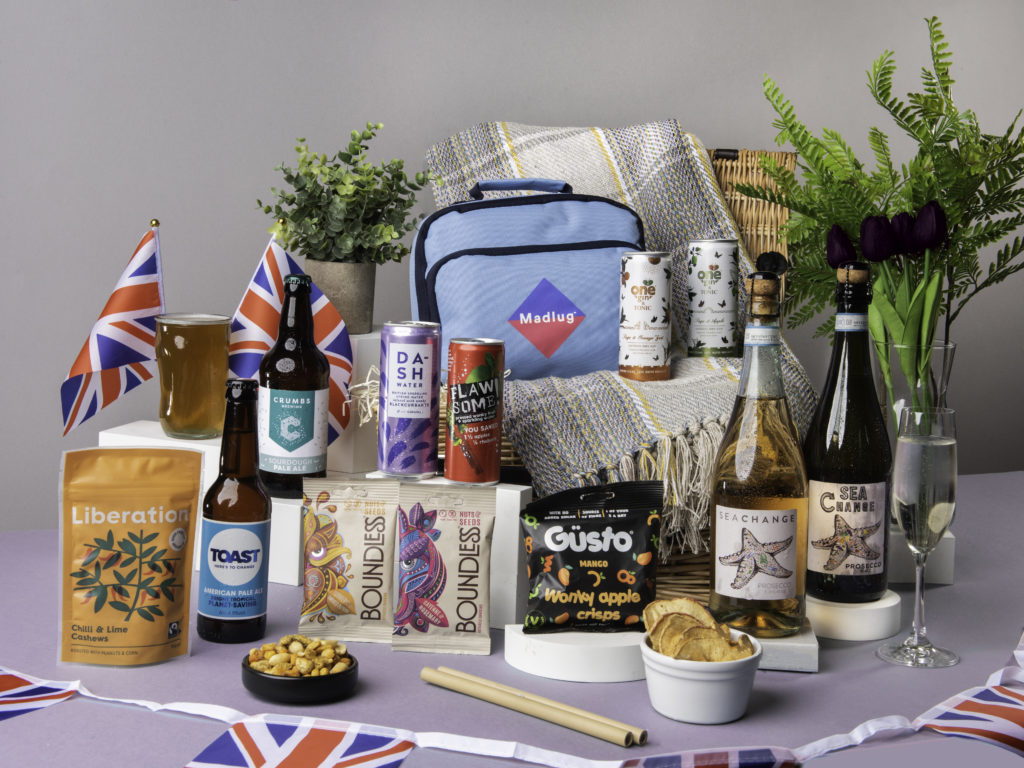 The Platinum Celebration Picnic Hamper, surrounded by its content including G&T cans from One Gin, beers from Crumbs Brewing and Toast Ale, Sea Change Prosecco Rosé, snacks from Güsto, Liberation Nuts and Boundless Activated Snacking, as well as a cool bag from Madlug and soft drinks from Dash and Flawsome! There's Union Jack flags dotted around them too.