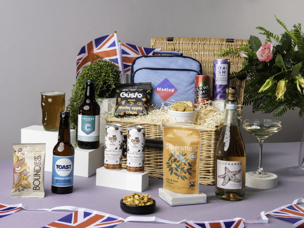 The Queen's Jubilee Picnic Hamper, surrounded by its content including G&T cans from One Gin, beers from Crumbs Brewing and Toast Ale, Sea Change Prosecco Rosé, snacks from Güsto, Liberation Nuts and Boundless Activated Snacking, as well as a cool bag from Madlug and soft drinks from Dash and Flawsome! There's Union Jack flags dotted around them too.