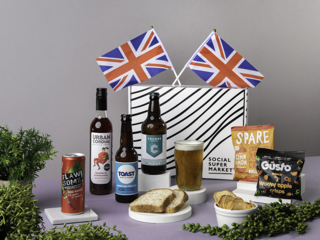 The Save Food Waste Jubilee Gift Box, surrounded by its content including beers from Crumbs Brewing and Toast Ale juice from Flawsome! and cordial from Urban Cordial and snacks from Spare and Güsto. They're on a picnic blanket and there's Union Jack flags dotted around them too.