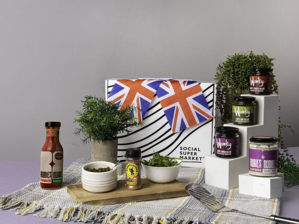 The Social Enterprise BBQ Booster Gift Box, surrounded by its content including Wonky Food Co relishes, Rubies in the Rubble tomato ketchup and plant-based mayo and the Rubbalicious Smoke 'n' Spice shaker. They're on a picnic blanket and there's Union Jack flags dotted around them too.