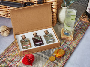 An Elephant Gin gift set of three mini gins presented on a recycled blanket with a ready-made Gin and Tonic cocktail next to the set with a circular cucumber slice inside the glass. 