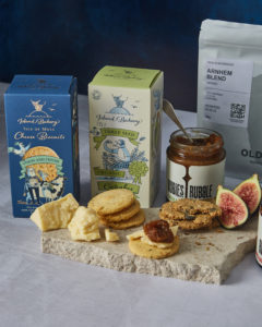 Two packs of Island Bakery biscuits with some of their biscuits in front of them on a naturally shaped marble board and some wedges of cheese, plus figs and a Rubies in the Rubble chutney jar with a spoon sticking out of the top.