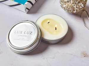 A LUX LUZ Letterbox Candle with its lid off and the two wicks lit. There's the corner of a Social Supermarket letterbox box in one corner and a sparkling bauble in the other.