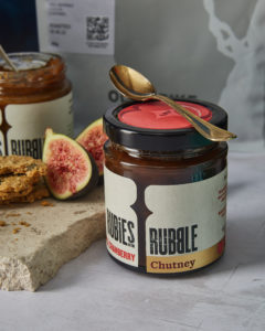 A jar of Rubies in the Rubble Apple and Cranberry chutney with a gold spoon on top, next to a naturally shaped board of marble with figs and other products in the background