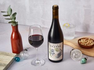 A bottle of Sea Change Organic Bobal wine with a glass poured next to it. In the background, a red vase with eucalyptus in and a cork stand with a sparkling drink, surrounded by baubles