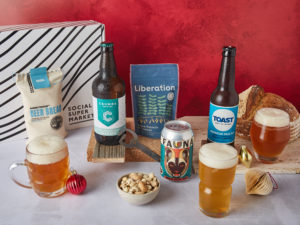 The Beer Lover Gift Box surrounded by the contents that are inside, including Barrett's Ridge Beer Bread Kit, Toast Ale and Crumbs Brewing beers in bottles and Fauna Brewing in a can, with glasses of the beers around them.