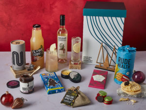 The Christmas Chill Gift Box surrounded by the contents that are inside, including Urban Cordial, Flawsome! Drinks apple juice, Tony's Chocolonely 51% Dark Chocolate Candy Cane, Rubies in the Rubble Chilli Onion Relish, a Vegan Bunny Christmas Tree-scented candle and more.