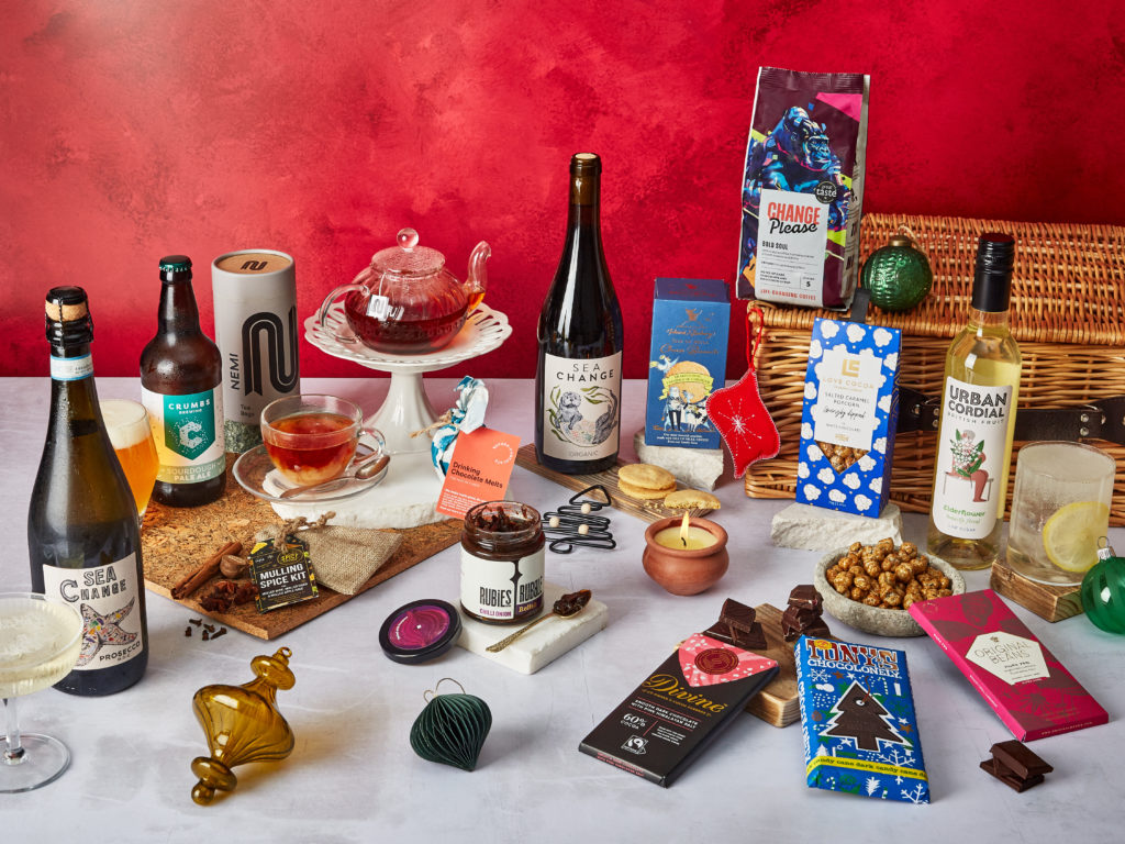 The Christmas Season Hamper with its products surrounding the wicker basket they come in, including Sea Change wines, Love Cocoa white chocolate popcorn, Change Please coffee, Urban Cordial, chocolate bars from Original Beans, Tony's Chocolonely and Divine Chocolate and more.
