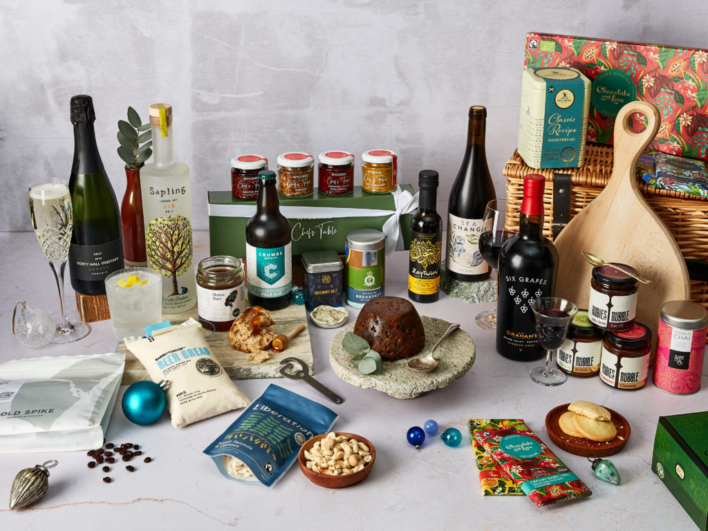 The Foodie Flavours Hamper with its products surrounding the wicker basket they come in, including Sea Change wines, Graham's Six Grapes Port, Wycombe Chef's Table Spice Gift Set, Zaytoun CiC Olive Oil, Rare Tea Company teas, Sapling Gin, Forty Hall Vineyards Sparkling Brut and more.