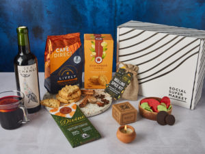 The Ginger & Spice Gift Box surrounded by the contents that are inside, including a half bottle of Sea Change Malbec wine, Cafédirect coffee, Divine Chocolate, a Dalit candle and more.