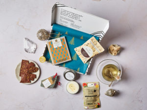 The Life's Little Luxuries Letterbox Gift by Social Supermarket – the box lid is open showing the Christmas tree tissue paper and its contents are scattered, including a Love Cocoa chocolate bar, a letterbox candle by LUX LUZ, tea by Tea People, a small bag of Nourish macaroons and a handmade Christmas decoration by Pivot.