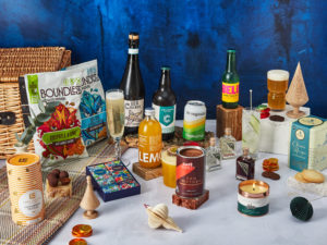 The Share & Unwind Hamper surrounded by the contents that are inside, including Sea Change Prosecco, Elephant Gin Tasting Trio, Cafédirect San Cristobal hot chocolate, Belu Tonic, Brewgooder Beer, a blanket, Chocolate and Love chocolate gift set, Vegan Bunny Christmas tree scented candle and more.