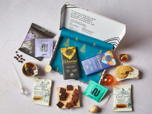 The Tea Break Letterbox Gift by Social Supermarket – the box lid is open showing the Christmas tree tissue paper and its contents are scattered, including Divine Chocolate bars, NEMI Teas sachets of teas, Island Bakery biscuit packs and Cafédirect coffee.