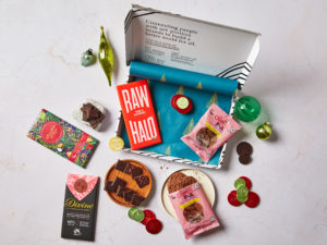 The Vegan Chocolate Letterbox Gift by Social Supermarket – the box lid is open showing the Christmas tree tissue paper and its contents are scattered, including chocolate bars from Raw Halo, Divine Chocolate and Chocolate and Love, plus Sweet FA biscuit packs.