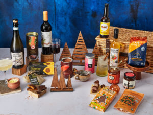 The Winter Favourites Christmas Hamper surrounded by the contents that are inside, including Symington Family Estates Altano wine, Toast Ale beer, Urban Cordial, Cafédirect coffee, Roots and Wings marmalade, Rubies in the Rubble relish, Tony's Chocolonely chocolate and more.