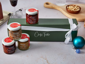 A dark green gift box with white writing "Wycombe Chef's Table" and a white ribbon surrounded by spice blends by Wycombe Chef's Table. In the background there's a glass of wine and a cheese board.
