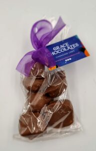 A bag of oat milk cinder toffee chocolates tied with a purple ribbon and Grace Chocolates Changing Lives tag