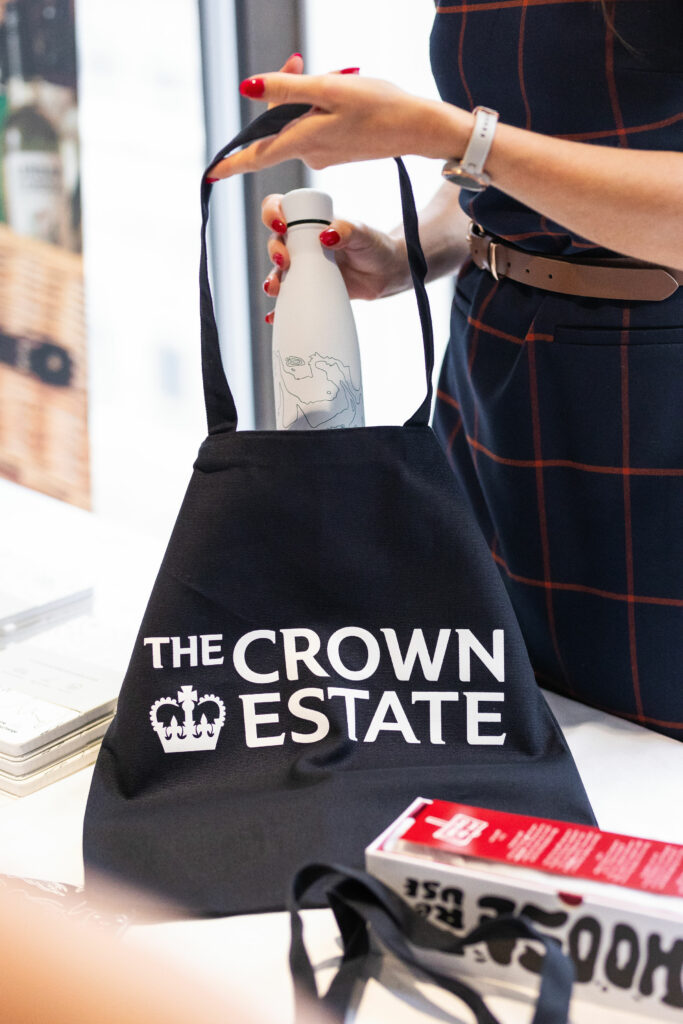 A woman puts a reusable bottle in a Crown Estate branded black tote bag